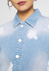 Světlomodrá džínová košile s knoflíkovým zapínáním, špičatým límečkem a rozptýlenými bílými vzory připomínajícími mraky. Hladká textura se stříbrnými knoflíky.