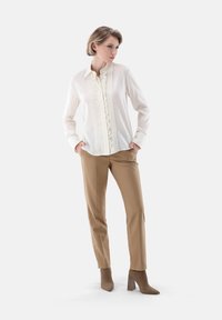 White blouse with a ruffled front and button closure, paired with beige trousers and taupe heeled ankle boots. Smooth fabric and tailored fit.