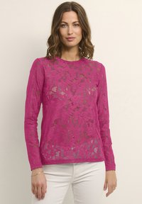 Top a maniche lunghe di pizzo rosa brillante con motivi floreali e texture trasparente, abbinato a pantaloni bianchi. Scollo rotondo semplice.