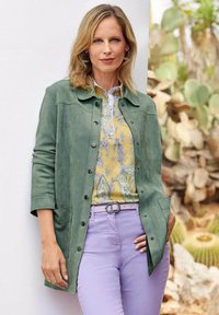 Groene suède jas met drukknoopsluiting, gecombineerd met een geel patroon blouse, licht paarse broek en een zilveren riem. Natuurlijke achtergrond.