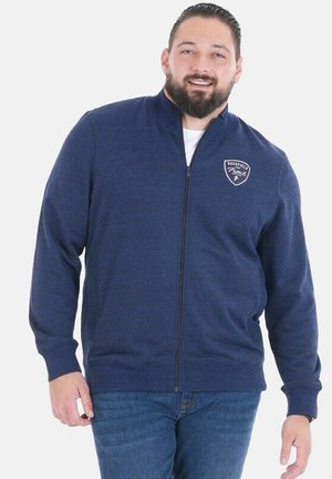 Homme avec une barbe portant une veste zippée bleu marine et un jean bleu, souriant devant un fond blanc uni.