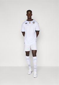 Hvit fotballuniform med svarte detaljer, med rund hals, korte ermer, matchende shorts og hvite knestrømper med mønstre.