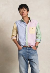 Chemise à rayures multicolores avec des sections bleu pâle, jaune, rose et verte ; comprend une poche jaune. Portée avec un jean bleu clair.