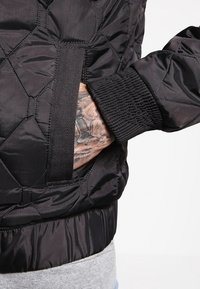 Svart vadderad jacka med ribbad mudd och detalj med sidoficka, med en slät, glänsande yta och kontrasterande texturer.