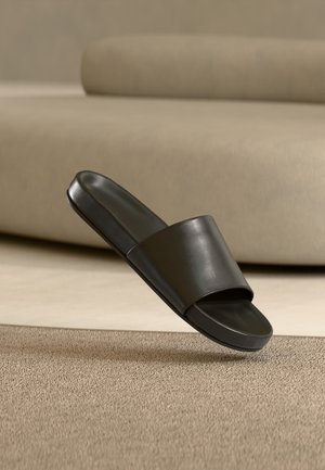 Schwarze Lederslide-Sandale, die über einem beigen strukturierten Teppich mit einem taupefarbenen Sofa im Hintergrund schwebt.
