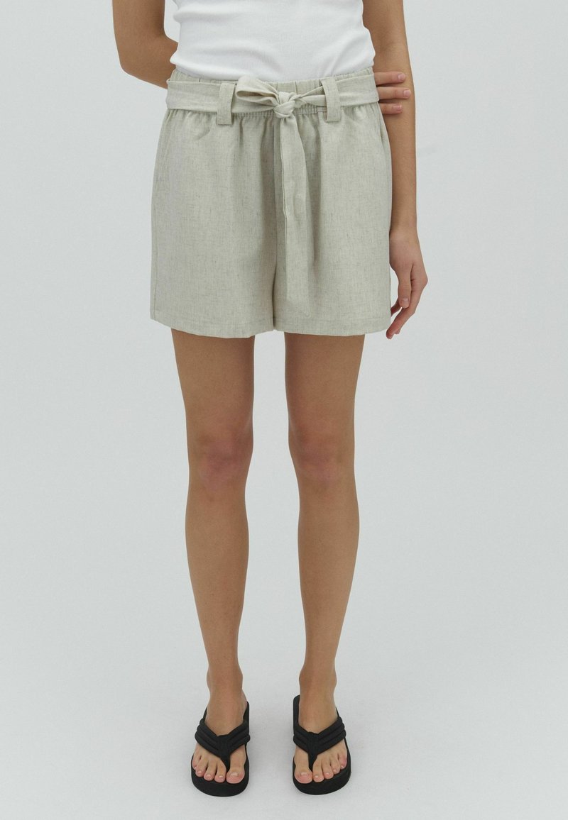 Femme portant un short beige taille haute avec ceinture nouée, un top blanc sans manches et des tongs noires, debout devant un fond uni.