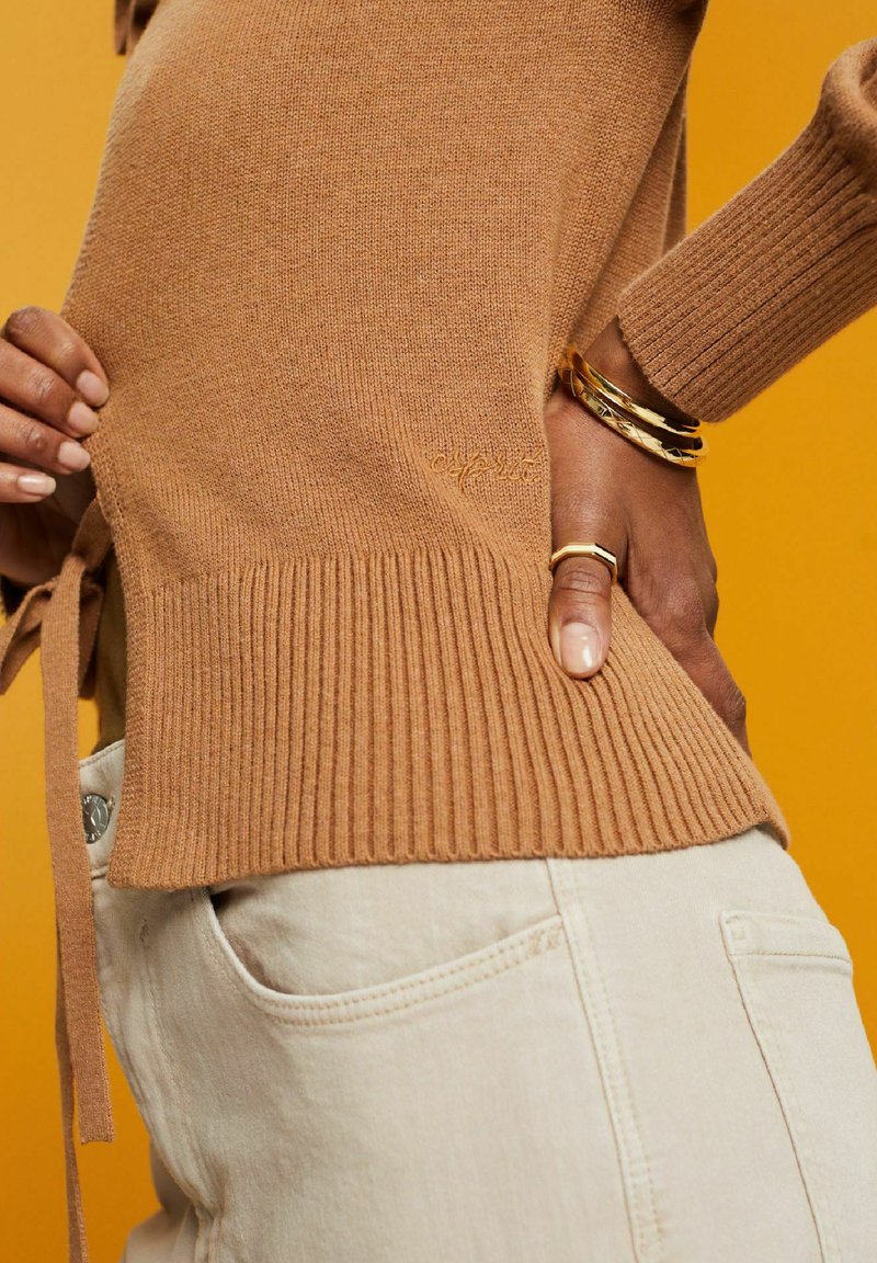 Pull en maille beige avec un ourlet côtelé et un détail de lien sur le côté, associé à un pantalon crème, le tout sur un fond jaune.