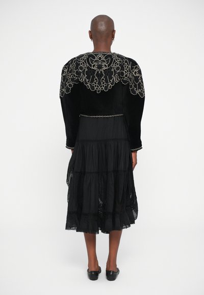 Black velvet jacket with intricate silver embroidery on the back, paired with a flowing black tiered dress and flat black shoes.