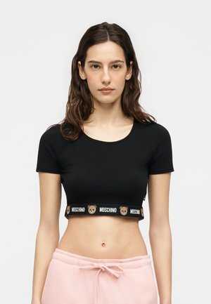 Młoda kobieta z długimi ciemnymi włosami, ubrana w czarny krótki top z pluszowym misiem i logo "MOSCHINO" oraz jasnoróżowe spodnie ze ściągaczem.