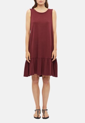 Femme portant une robe bordeaux sans manches avec un ourlet volanté, debout, les mains détendues le long du corps, et des sandales noires à brides en T sur fond blanc.
