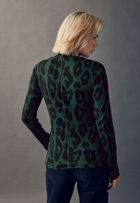Zöld hosszú ujjú felső fekete leopárdmintával, bordázott textúrával és a háton végigfutó varrási részlettel. Sötét farmernadrággal párosítva.