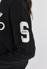 Czarna bluza z okrągłym dekoltem, z białą welurową łatką "S" na lewym rękawie oraz fakturowanym dzianinowym materiałem.
