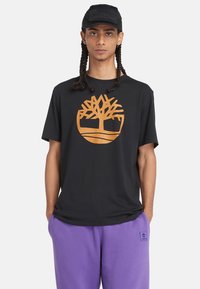 Schwarzes Baumwoll-T-Shirt mit großem orangefarbenem Timberland-Baumlogo. Getragen mit lila Trainingshose, die einen entspannten Schnitt und kurze Ärmel hat.