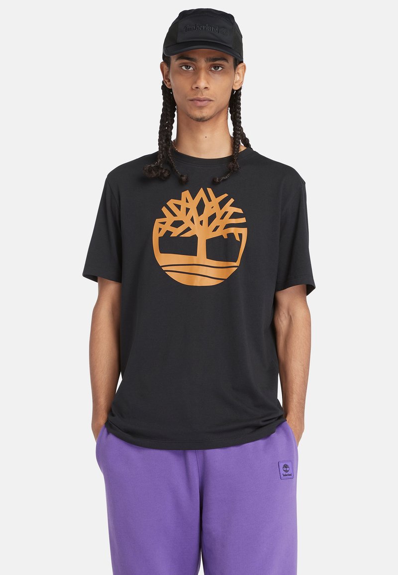Schwarzes Baumwoll-T-Shirt mit großem orangefarbenem Timberland-Baumlogo. Getragen mit lila Trainingshose, die einen entspannten Schnitt und kurze Ärmel hat.