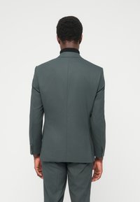 Σακάκι κουστουμιού προσαρμοσμένης ραφής, σκούρο πράσινο, μονόκουμπο με δύο κουμπιά, υφασμάτινη υφή, γιακάς με αιχμές και κοντράστ μαύρο γιακά.