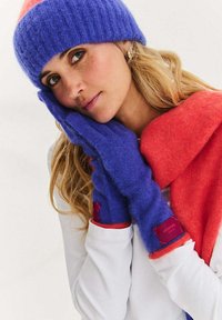 Pletená čepice ve fialové a růžové barvě, ladící fialové bezprsté rukavice a výrazná červená šála s jemnou texturou a kontrastními manžetami.