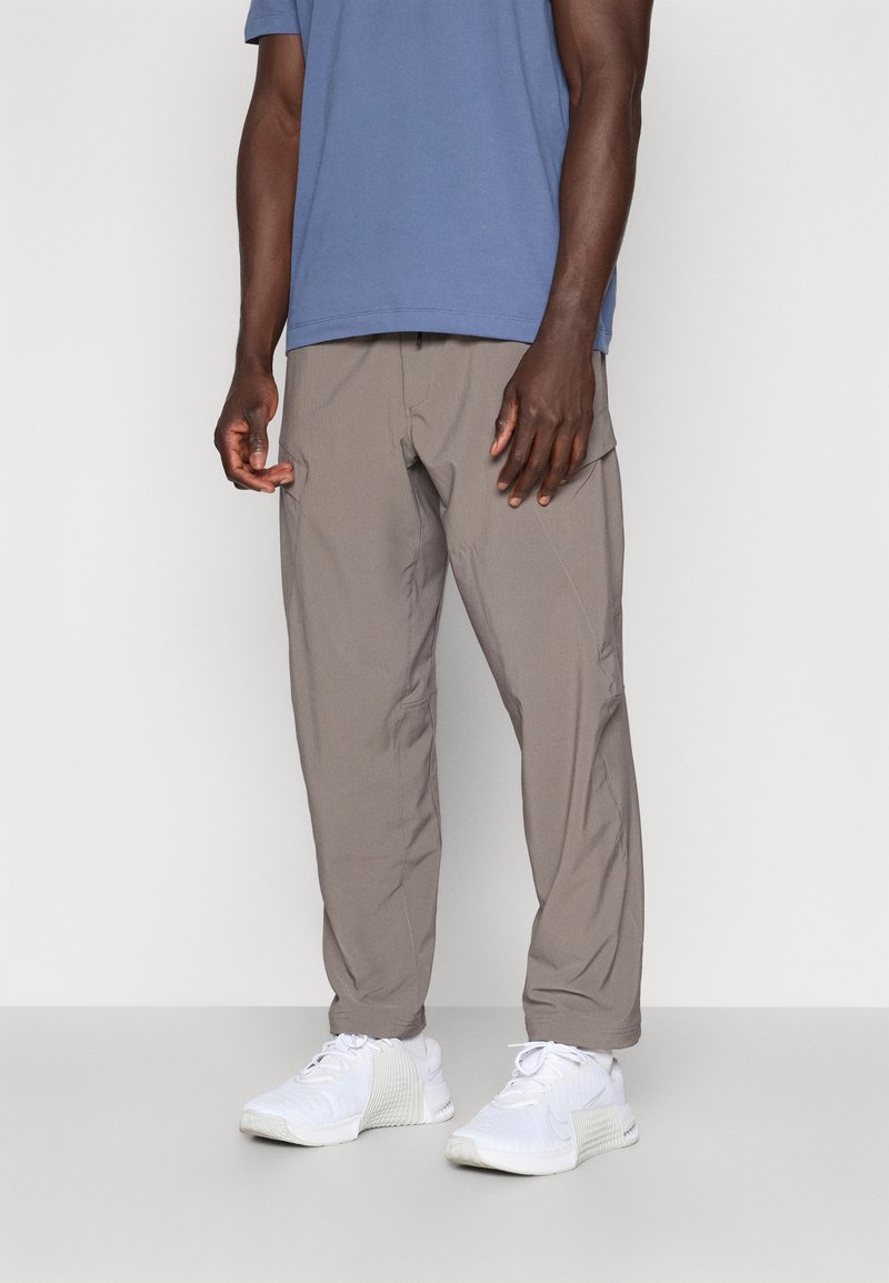 Pantaloni ușori, gri, cu croială slim-fit, prevăzuți cu buzunare laterale; asortați cu o cămașă albastră și pantofi sport albi. Detalii de cusătură vizibile și linii curate.