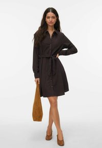 Mustast nööpidega särk-kleit pikkade varrukatega, vöökohal vöö ja helkiva seelikuga. Modell hoiab käes beežikõrvenahast kotti ning kannab pruune kontsakingi.