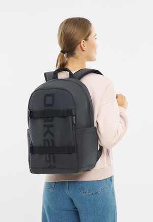 Black backpack with large logo, adjustable straps, side pockets, and a textured finish. Worn over a pink top with light blue jeans.