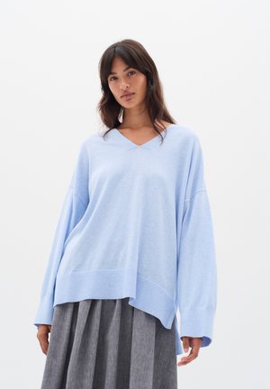 Hellblaues Übergrößen-Sweatshirt mit V-Ausschnitt, langen Ärmeln und geripptem Saum. Softtextur, lässige Passform, getragen über einem dunkelgrauen Faltenrock.