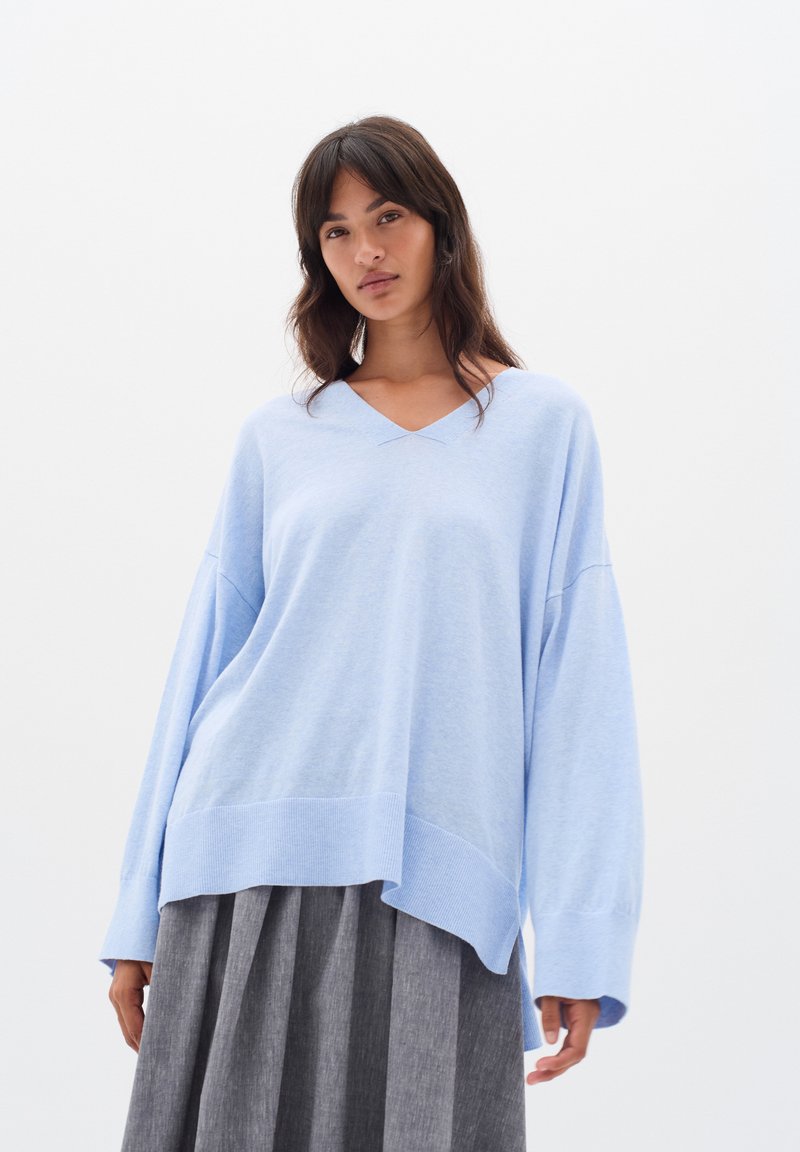 Hellblaues Übergrößen-Sweatshirt mit V-Ausschnitt, langen Ärmeln und geripptem Saum. Softtextur, lässige Passform, getragen über einem dunkelgrauen Faltenrock.