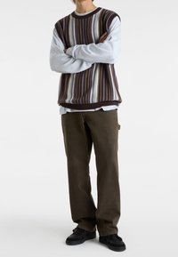 Den stickade tröjan har horisontella ränder i brunt, krämfärgat och lila, och bärs tillsammans med en grå långärmad skjorta och bruna byxor. Svarta sneakers.