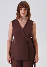 Donna con capelli castani mossi fino alle spalle, che indossa un top senza maniche marrone avvolgente con fibbia laterale e pantaloni abbinati, davanti a uno sfondo neutro.