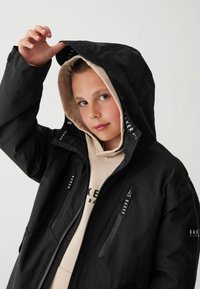 Schwarze, wasserdichte Jacke mit Kapuze, ausgestattet mit Reißverschlusstaschen und Logo-Details, getragen über einem beigen Hoodie. Glatte Textur, modernes Design.