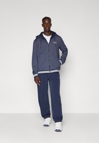 Camisola de capuz azul-marinho com riscas brancas nas mangas e na bainha, combinada com calças de jogging a condizer. O conjunto apresenta um logótipo no peito e na perna.