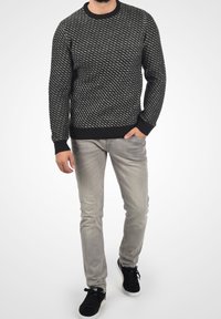 Uomo che indossa un maglione a righe bianco e nero, jeans grigio chiaro e sneaker nere, in piedi con una mano in tasca.