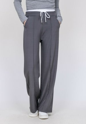 Personne portant un pantalon gris à jambes larges avec un cordon blanc et des baskets blanches, les mains dans les poches, sur un fond uni.