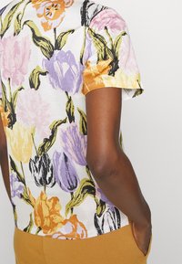 Camiseta de manga corta con estampado floral que presenta una base de tela blanca con impresiones de tulipanes multicolores en amarillo, púrpura y naranja, con un diseño de cuello redondo.