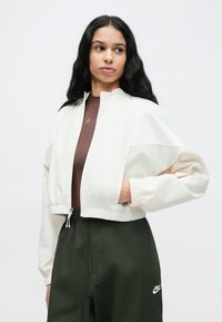 Λευκό και κρεμ μπουφάν σε μπλοκ χρωμάτων, κοντό, με ψηλό γιακά και ελαστικά μανίκια, σε συνδυασμό με καφέ τοπ και σκούρο πράσινο παντελόνι με λογότυπο.