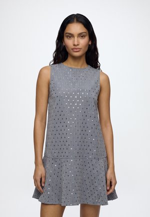 Femme aux longs cheveux foncés portant une robe grise sans manches décorée de lignes verticales de petites paillettes brillantes sur un fond clair uni.