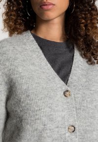 Szary wełniany kardigan z dekoltem w serek, z dwiema guzikami. Miękka tekstura i subtelny wzór na całej powierzchni. Noszony na ciemną koszulę.