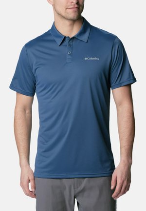 Uomo con polo blu a maniche corte Columbia e pantaloni grigi, in piedi davanti a uno sfondo bianco uniforme.