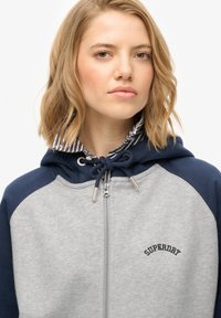 Sudadera con cremallera gris con acentos en azul marino, capucha con cordón y forro a rayas. Presenta un logotipo bordado de "SUPERDRY" en negro.