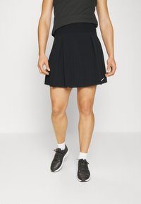 Musta plisattu urheiluhame, jossa on leveä vyötärönauha, valmistettu kevyestä kankaasta. Yhdistetty mustiin tennareihin ja valkoisiin sukkiin, neutraalilla taustalla.