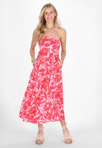 Rochie maxi fără bretele, de culoare roz, cu un model floral roșu, având butoane pe față și buzunare, asortată cu espadrile de culoare deschisă.