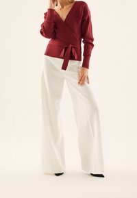 Donna che indossa un maglione a V rosso con fiocco e pantaloni bianchi a vita alta e largo, in piedi con una mano vicino al collo e l'altra sulla coscia.