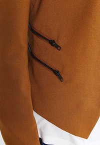 Ruskea teksturoitu takki, jossa kaksi mustaa vetoketjullista taskua sivuilla. Helmassa erottuva saumakuviointi ja alla kontrastia tuova valkoinen kangas.