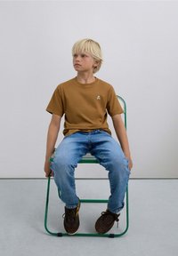 Brown cotton t-shirt with a small skull graphic, blue denim jeans, and dark sneakers, seated on a green metal chair against a light backdrop.