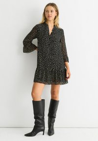 Black dress with polka dots, featuring a collar, long bell sleeves, and a flared hem. Paired with knee-high black leather boots.