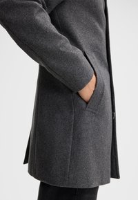 Grauer Wollmantel mit strukturiertem Finish, ausgestattet mit einer Seitentasche und einer geraden Silhouette. Der Kragen ist aus diesem Winkel nicht sichtbar.