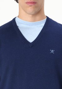 Tummansininen V-pääntie neule, jossa on sileä pinta ja koristereunukset, käytetty vaaleansinisen pyöreäkaulaisten T-paidan päällä. Rinnassa pieni brodeerattu logo.