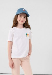 Witte t-shirt met korte mouwen en een kleurrijk logo op de borst, gecombineerd met lichtroze sweatpants. Een blauwe pet met een logo wordt gedragen.