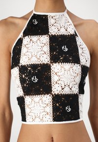 Top corto all'uncinetto con motivi a quadri alternati in bianco e nero, caratterizzato da intricati design in pizzo e un colletto alto. Dettaglio del logo ricamato.