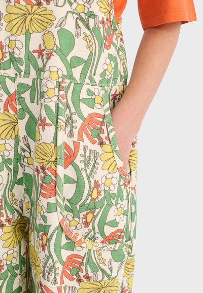 Main dans la poche, portant un pantalon à imprimé floral coloré en jaune, vert, blanc et orange, accompagné d'un haut à manches courtes orange.