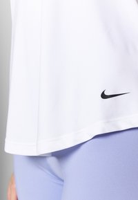 T-shirt de sport blanc avec un ourlet arrondi et un logo Nike noir en bas à gauche, associé à des leggings lavande clair, texture de tissu lisse.