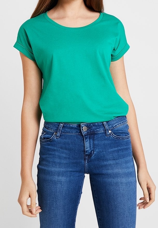 Femme portant un t-shirt vert uni à manches courtes, rentré dans un jean bleu taille haute avec les manches retroussées.